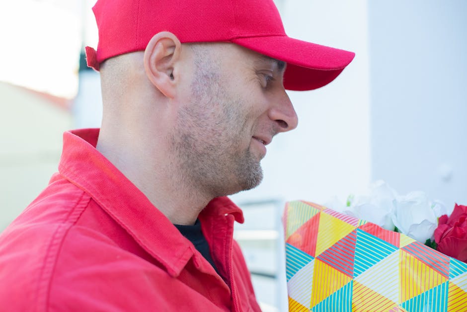 A man wearing a red cap and red jacket is holding a vibrant floral gift wrapped in colorful, geometric-patterned paper with white tissue paper peeking out, standing outdoors with a soft focus backgrou