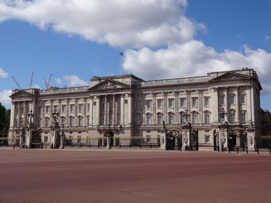 A large, historic stone building with classical architectural details, featuring tall windows, decorative columns, and ornate iron gates at the entrance, situated against a backdrop of blue sky with s