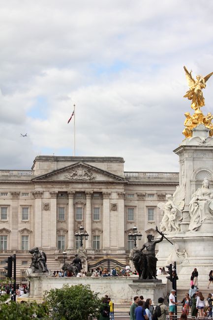 The image shows an outdoor scene with a historic building featuring classical architecture, tall columns, and a central pediment. In front of the building, there is a large ornate monument with a whit