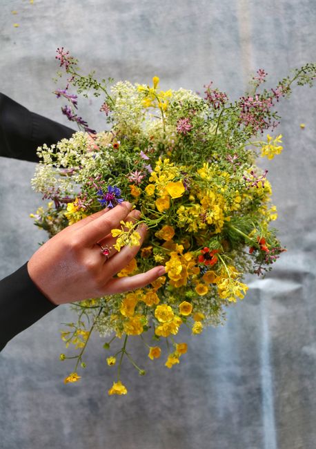 A hand holding a vibrant bouquet of fresh, wildflowers featuring yellow primroses, delicate white and purple blooms, small pink and red blossoms, and sprigs of greenery. The bouquet is loosely arrange